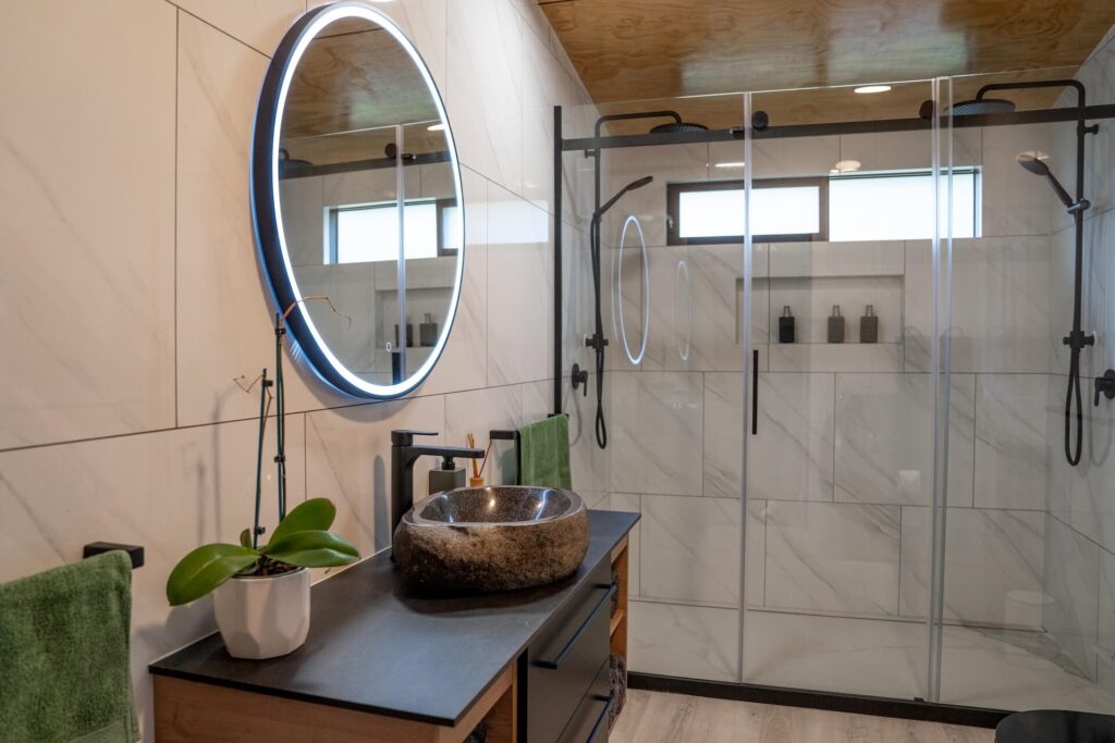 The Karamea-inspired modern bathroom features a large circular illuminated mirror, stone sink, and glass-enclosed shower. Plush towels hang on the wall, and a potted plant adds a touch of nature to the countertop.