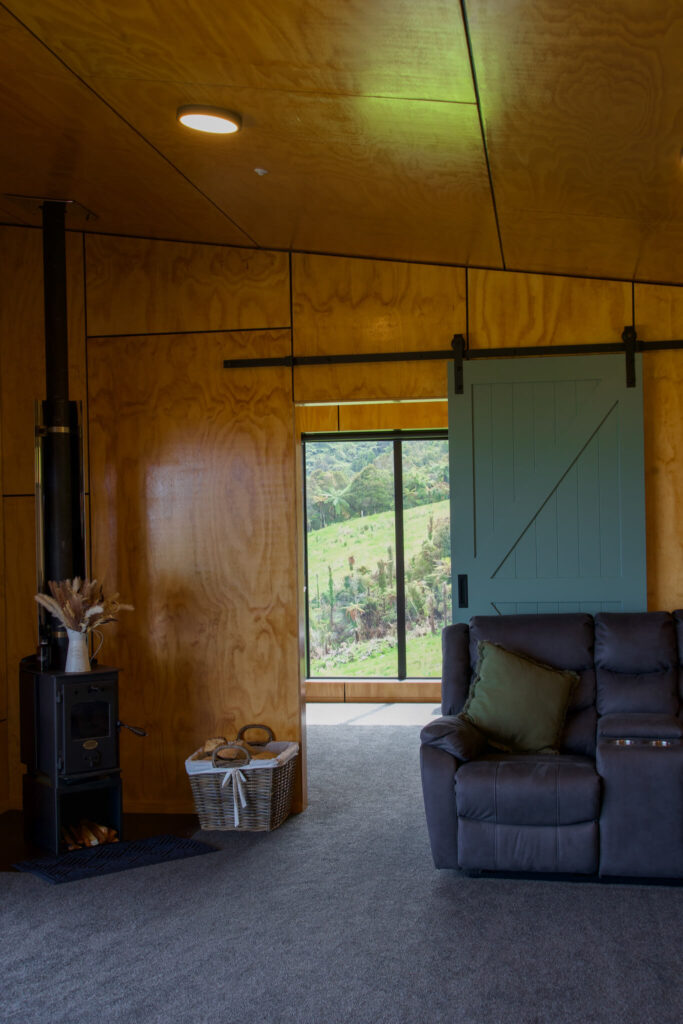 Cozy room with a wood-burning stove, a gray sofa, and a basket of firewood. A sliding barn door is ajar, revealing lush Karamea greenery through large windows.