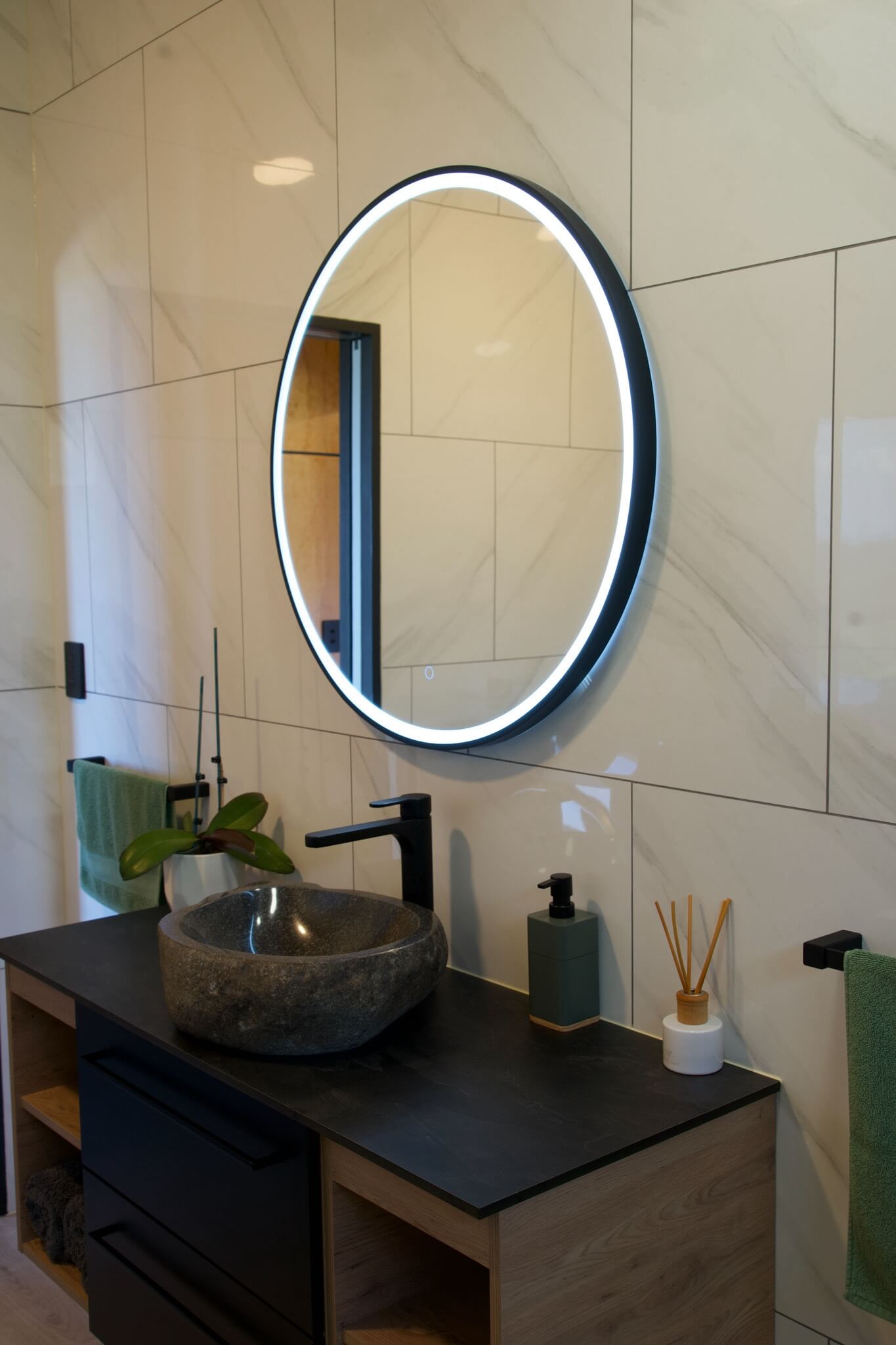 The Karamea-inspired bathroom features a round backlit mirror, a stone sink, and a sleek black faucet. The countertop is adorned with a stylish soap dispenser and diffuser, creating an ambiance of tranquil elegance.