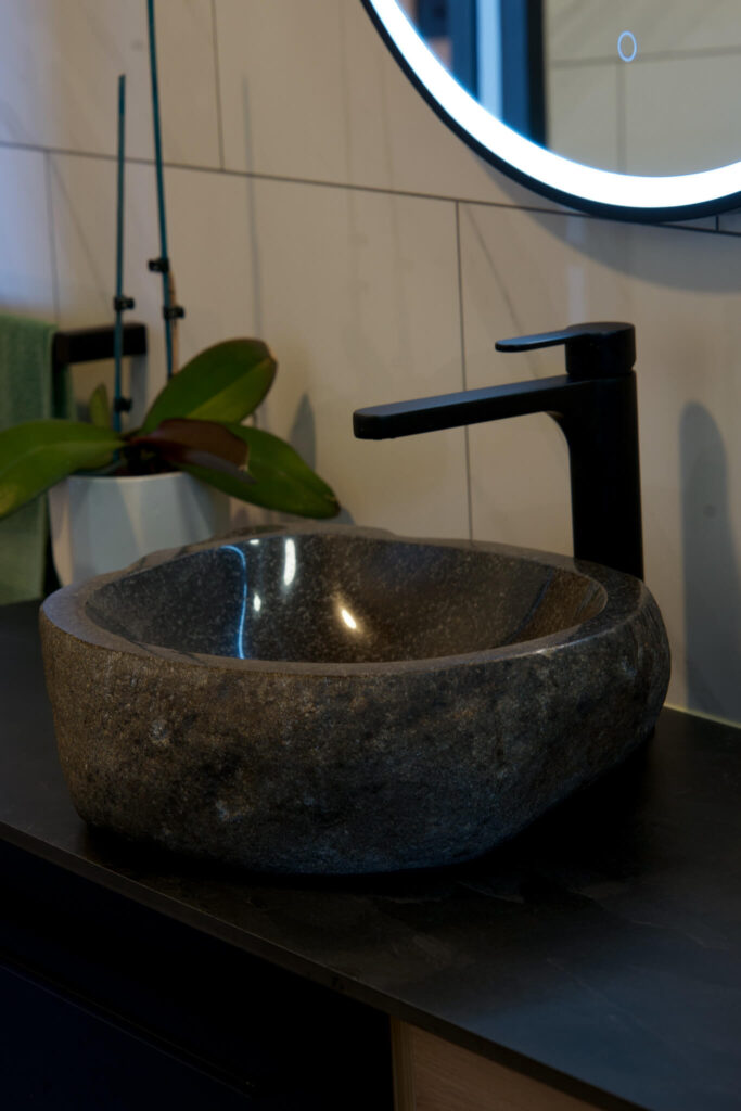 A stone sink with a modern black faucet is set against a tiled wall, echoing the serene elegance of Karamea. A round mirror with LED lighting hangs above, and a potted plant graces the counter.