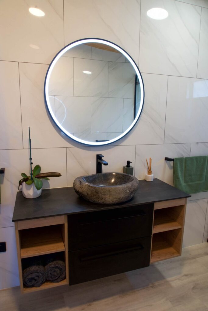 Modern bathroom with a round, illuminated mirror above a stone sink. A touch of Karamea charm is added with the potted plant and toiletries on the countertop. Open shelves below hold neatly stacked towels, enhancing the serene atmosphere.