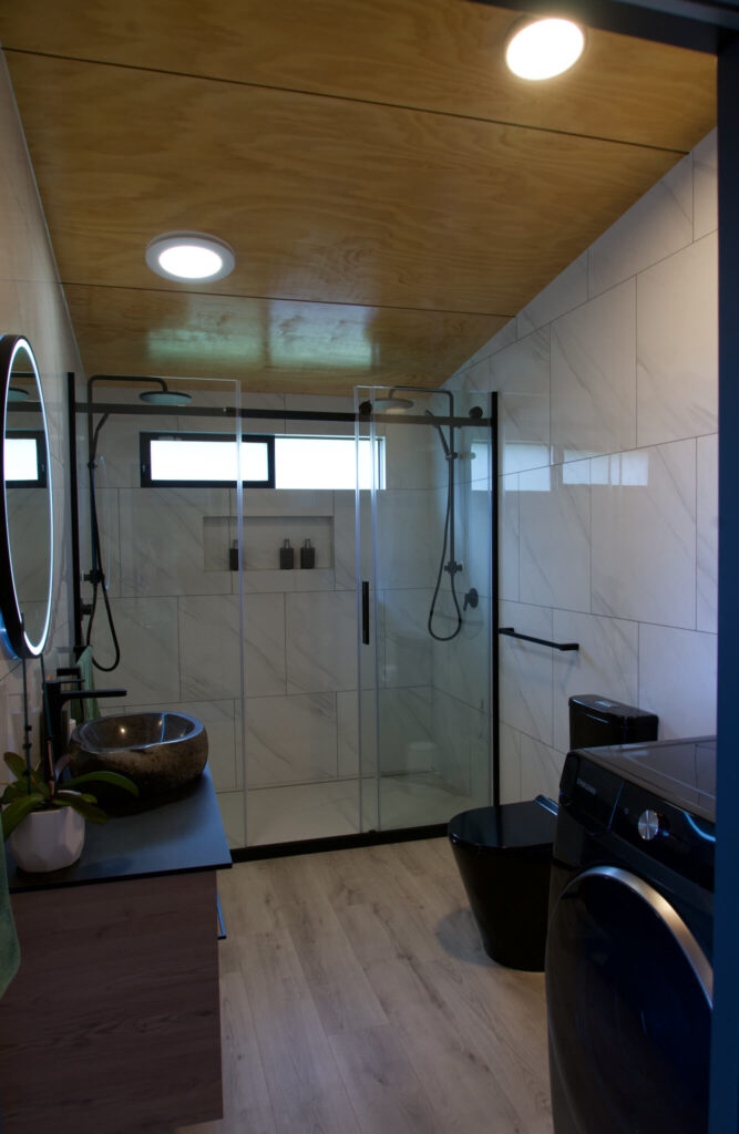 Discover a modern Karamea bathroom with a wood ceiling, marble walls, and glass shower. It features a sleek black sink, matching toilet, and washer. A circular mirror graces the space above the sink, while natural light pours through a small window positioned above the shower.