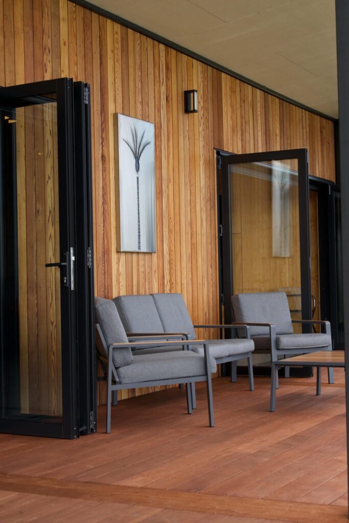 Two gray chairs rest on the wooden deck, beside glass patio doors and a wood-paneled wall reminiscent of Karamea's rustic charm. A rectangular black-and-white artwork adds a touch of elegance above.