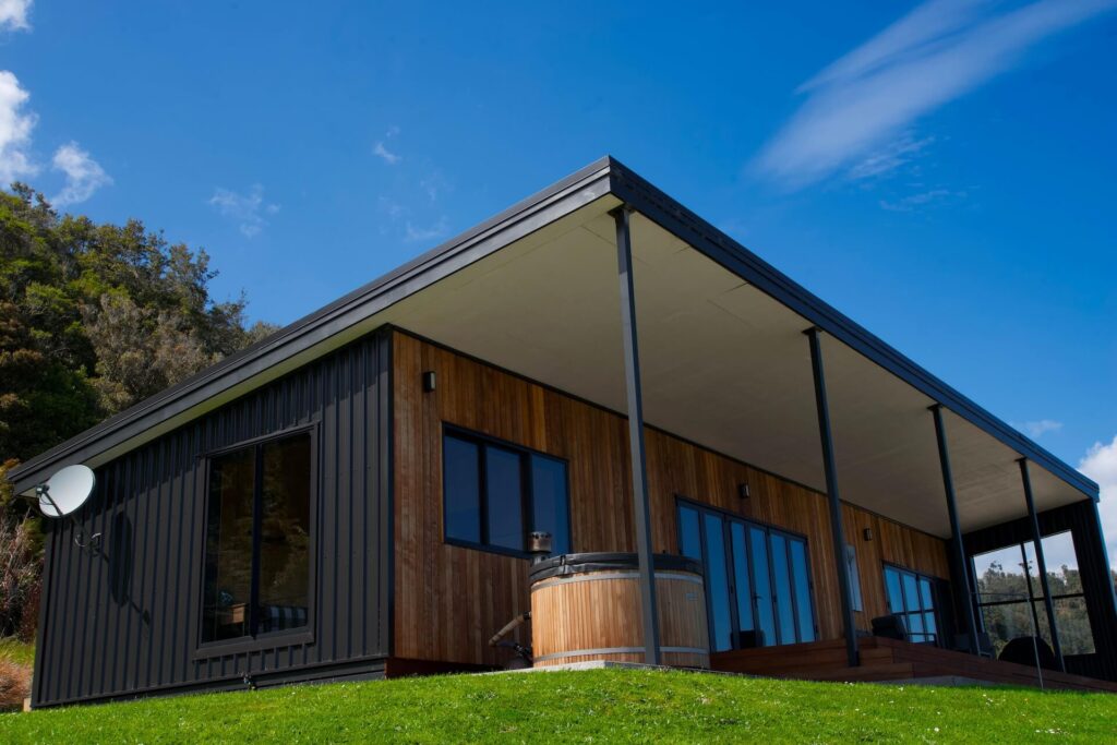 A modern house in Karamea showcases a wooden and black metal exterior, with large windows, a covered patio, and a hot tub nestled on a grassy slope under the expansive blue sky.