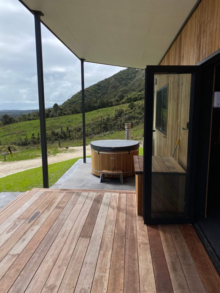 Outdoor patio with wooden decking and a circular hot tub, offering a slice of Karamea’s beauty. An open glass door leads inside. Enjoy the scenic view of green hills and a cloudy sky in the background.