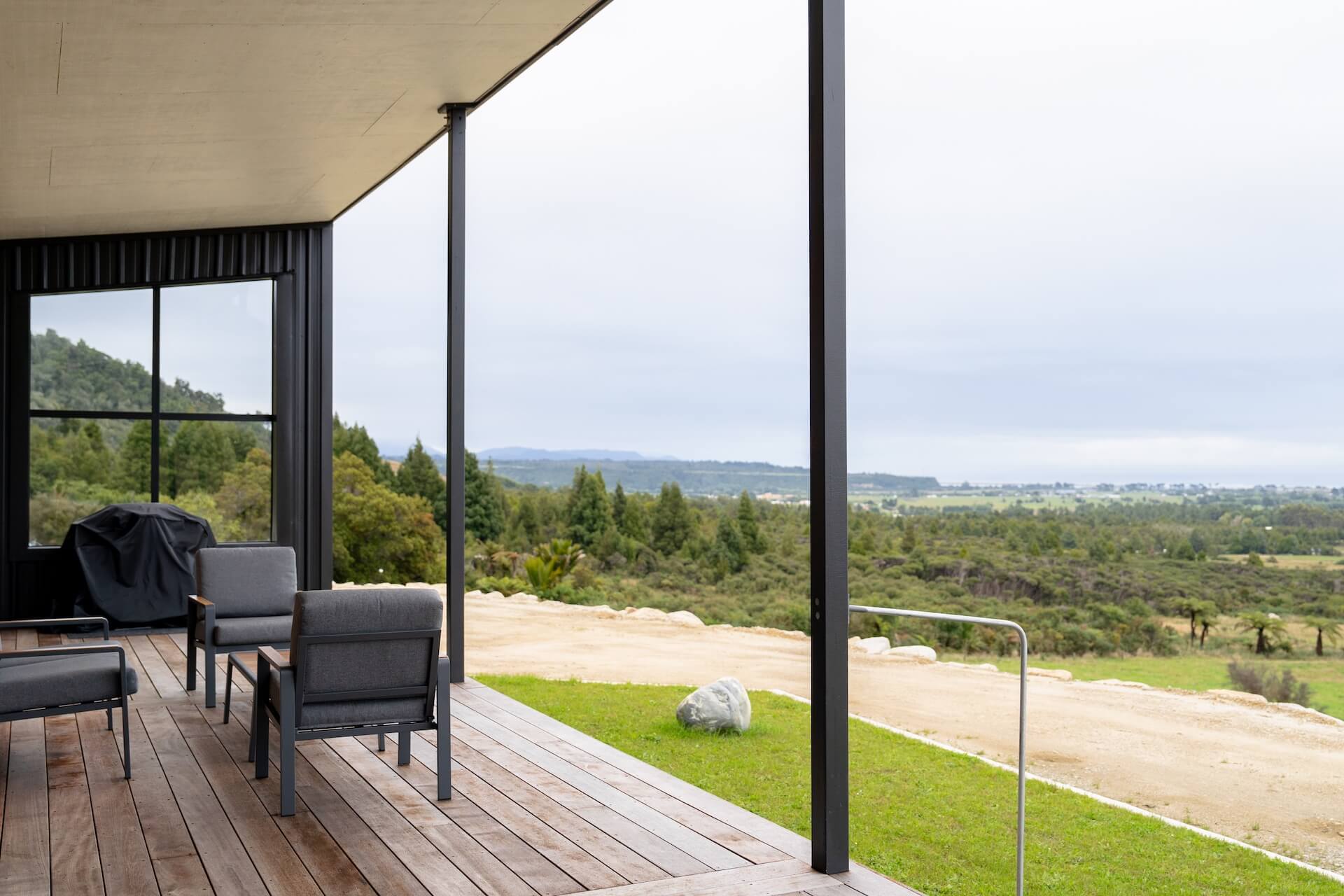 The modern patio with chairs overlooks a grassy landscape and forested area under a cloudy sky, reminiscent of the serene beauty found near Karamea.