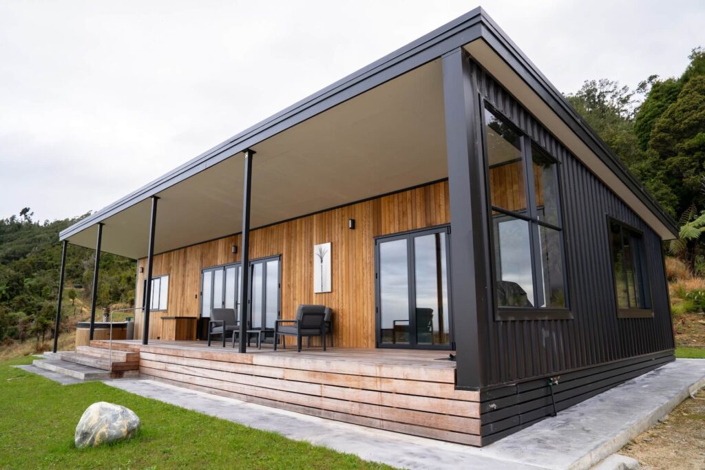 A modern house in Karamea with a rectangular shape, featuring a mix of wood and black metal siding, large windows, and a spacious porch with outdoor seating.