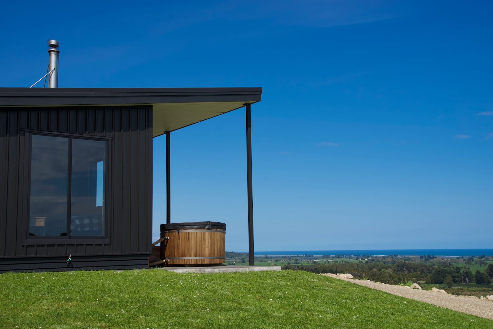 Contemporary black house with a veranda and hot tub in Karamea overlooks a grassy hill and distant sea under a clear blue sky.