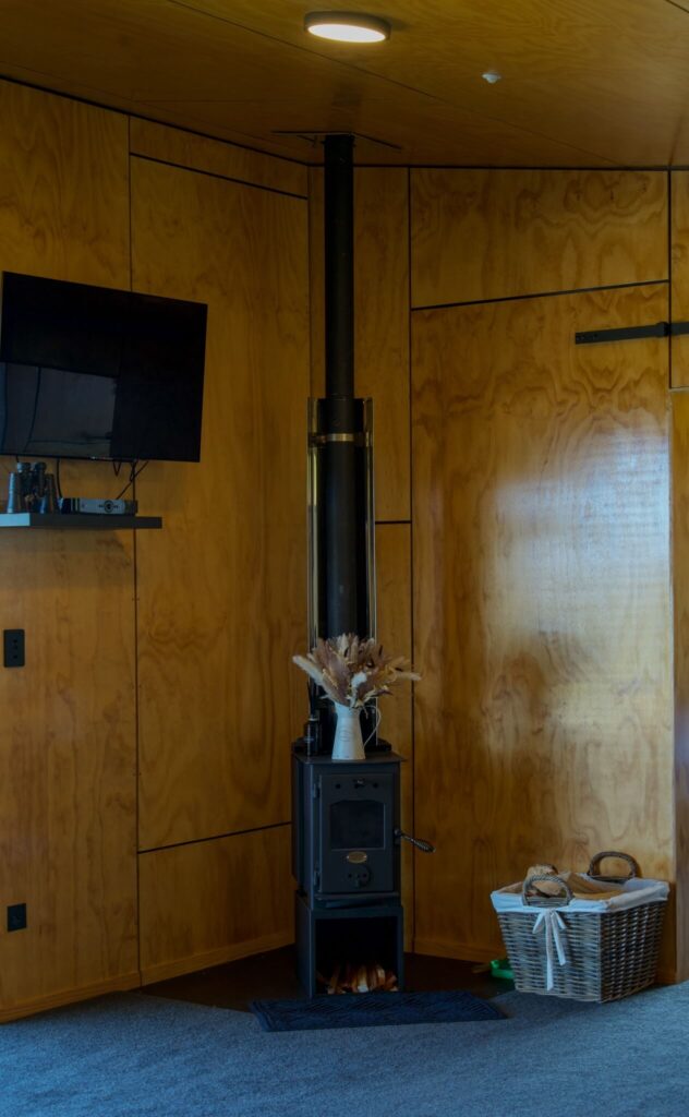 In the cozy Karamea wood-paneled room corner, a black wood-burning stove complements the wall-mounted TV. A wicker basket brimming with logs sits nearby, while a vase of dried flowers adds a touch of elegance atop the stove.