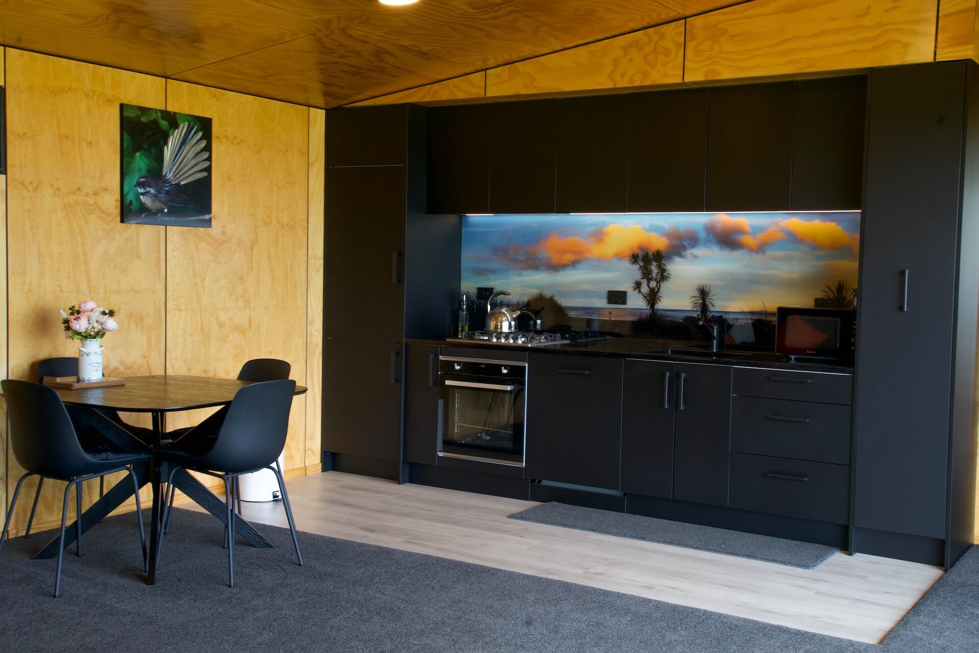 Modern kitchen with sleek black cabinets, a vibrant backsplash, and a dining table featuring black chairs. The wooden walls add warmth, while a floral arrangement graces the table. This Karamea-inspired space perfectly balances contemporary design with natural charm.