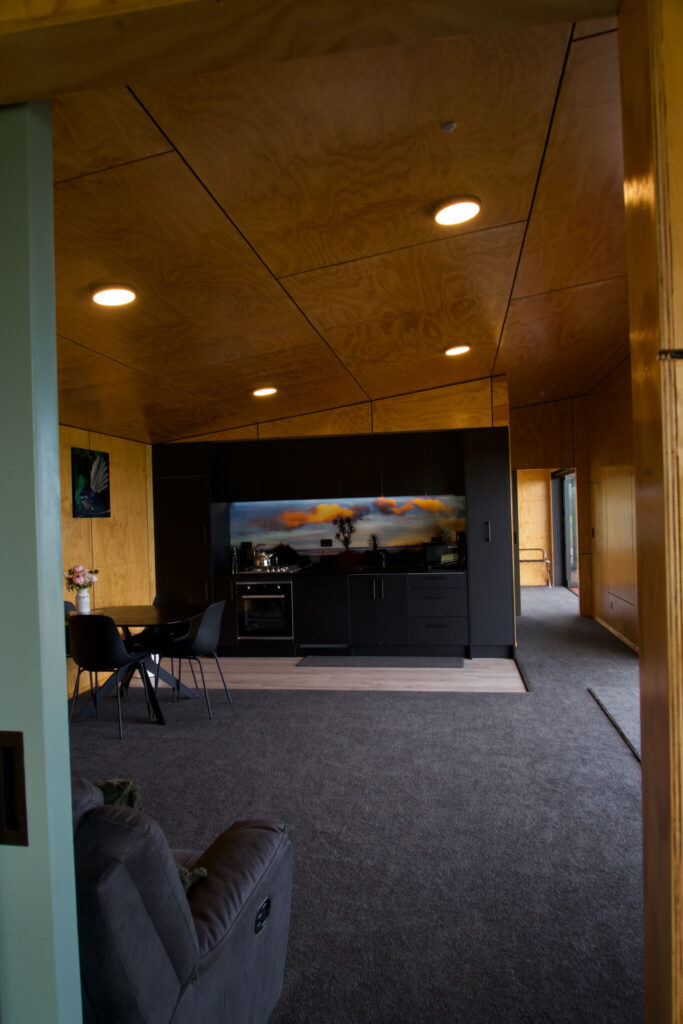 View of a modern living space in Karamea with wood-paneled walls, a kitchen area featuring black cabinets, a small dining table, and gray carpet. A large scenic artwork is displayed prominently in the kitchen area.