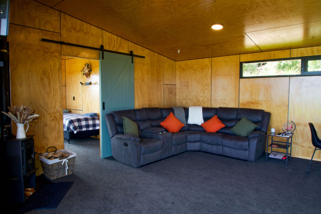 Cozy living room with a gray sectional sofa, orange and white pillows, wooden walls reminiscent of Karamea’s rustic charm, and a sliding door leading to a bedroom with a checkered bedspread.