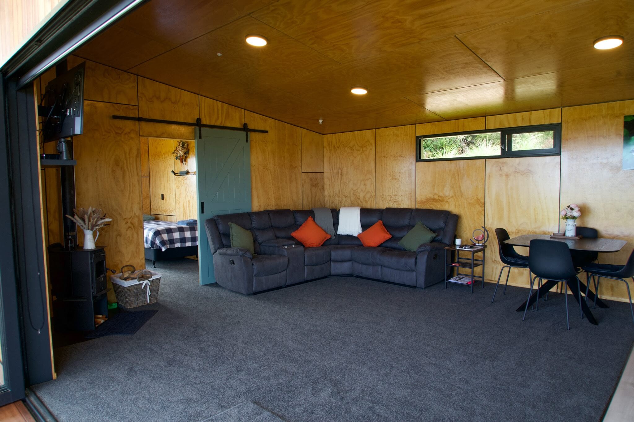 A stylish, modern living room in Karamea features wooden walls and a large gray sectional sofa adorned with colorful cushions. A sleek dining table with chairs complements the setup, offering a seamless view into the bedroom through a sliding door.