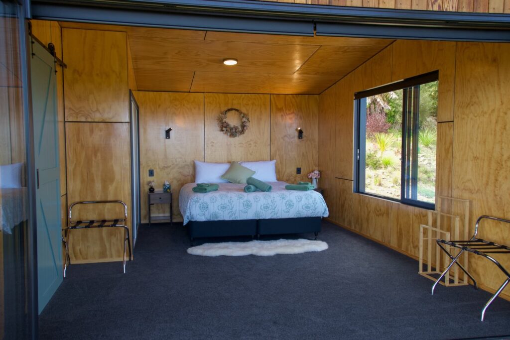 The Karamea-themed bedroom boasts wooden walls and a ceiling, a cozy double bed adorned with green and white bedding, bedside tables, and expansive windows. Two empty luggage racks await your arrival, alongside a decorative wreath that adds a charming touch.