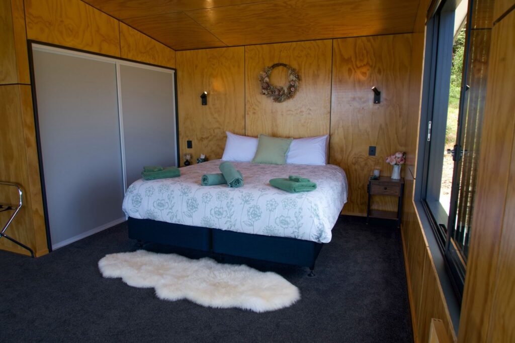 Cozy bedroom with wooden walls and a Karamea charm, featuring a bed with floral bedding and green towels. A small wreath hangs above the bed, resting on a fluffy rug over the gray carpeted floor, while a large window on the right invites in natural light.