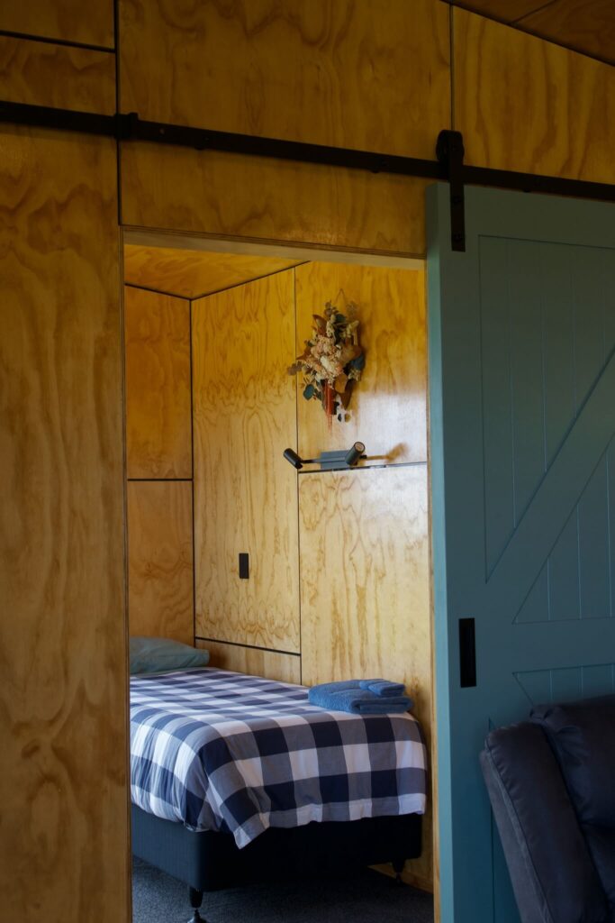 A room with wooden walls features a bed with a checkered blanket, a decorative wall piece inspired by Karamea, and a blue sliding door.