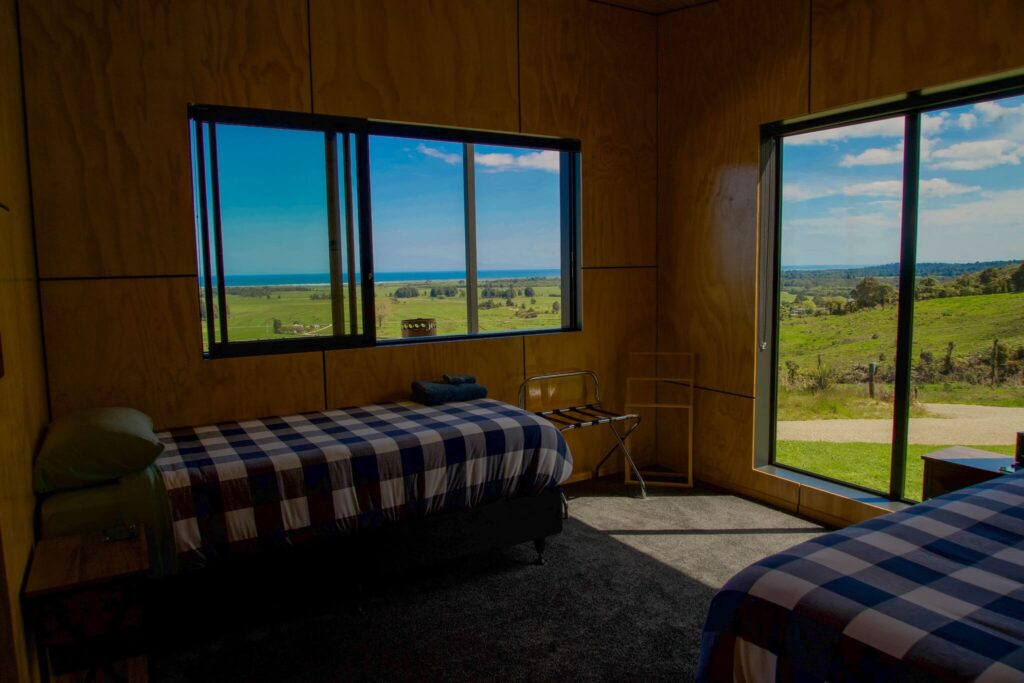 A room in Karamea with two single beds, blue checked bedding, and large windows showcasing a scenic view of green fields and the sea in the distance.