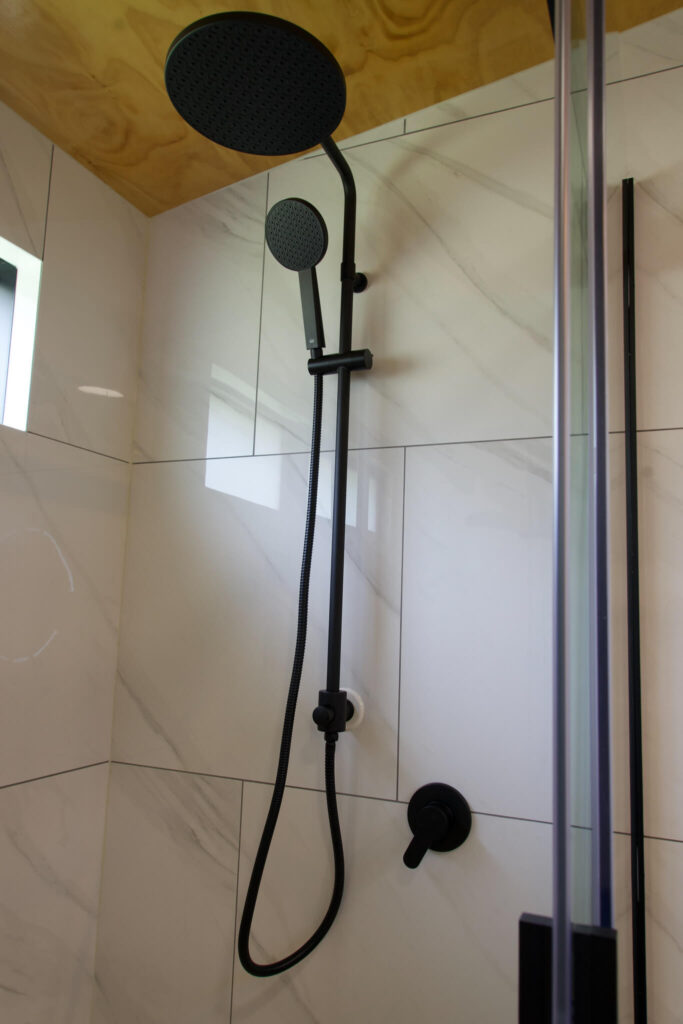 The modern Karamea shower boasts a matte black wall-mounted rainfall showerhead and handheld nozzle, beautifully contrasting against pristine white tiled walls.