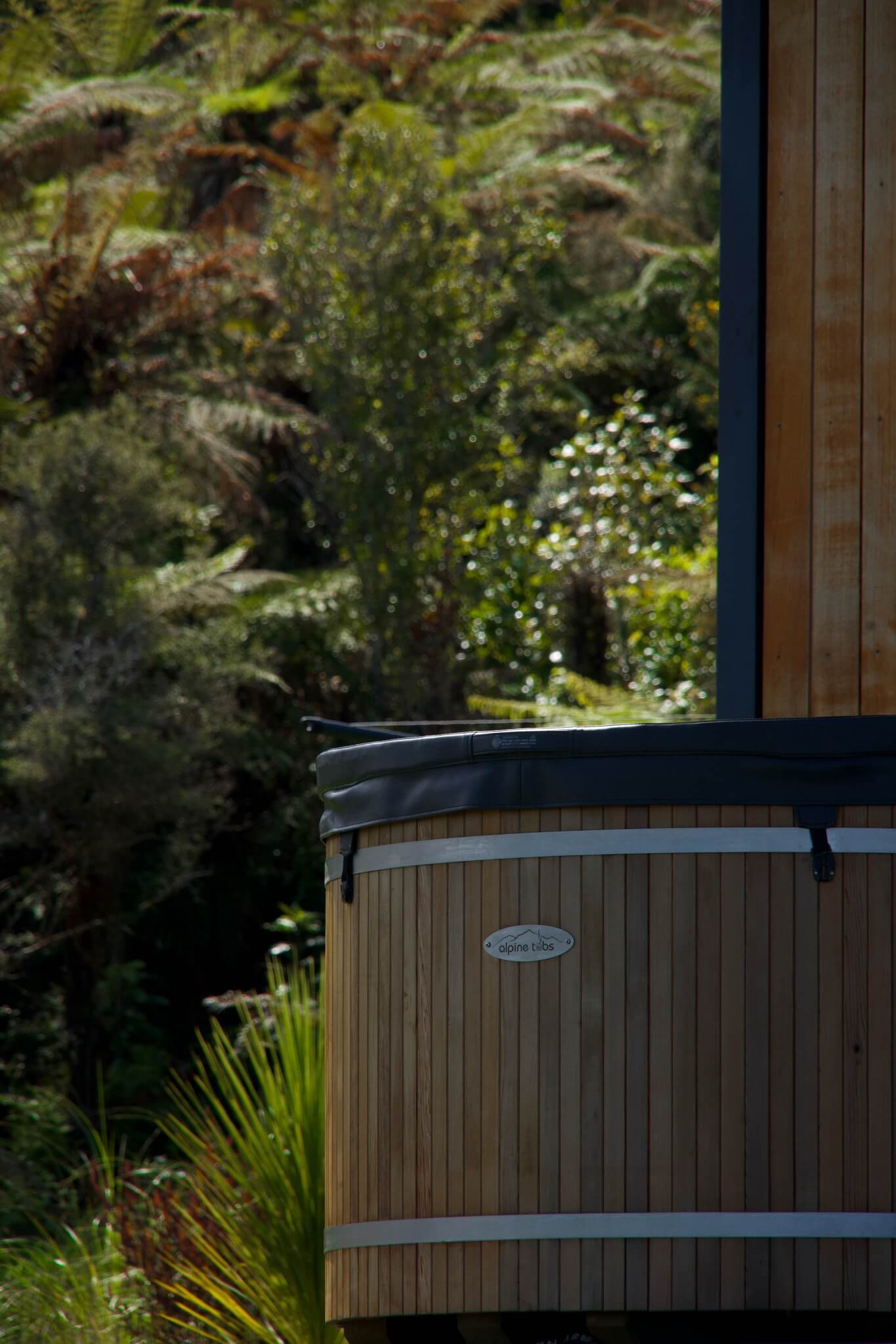 Nestled in the serene beauty of Karamea, a wooden hot tub rests against a backdrop of lush greenery and an elegantly crafted wooden structure.