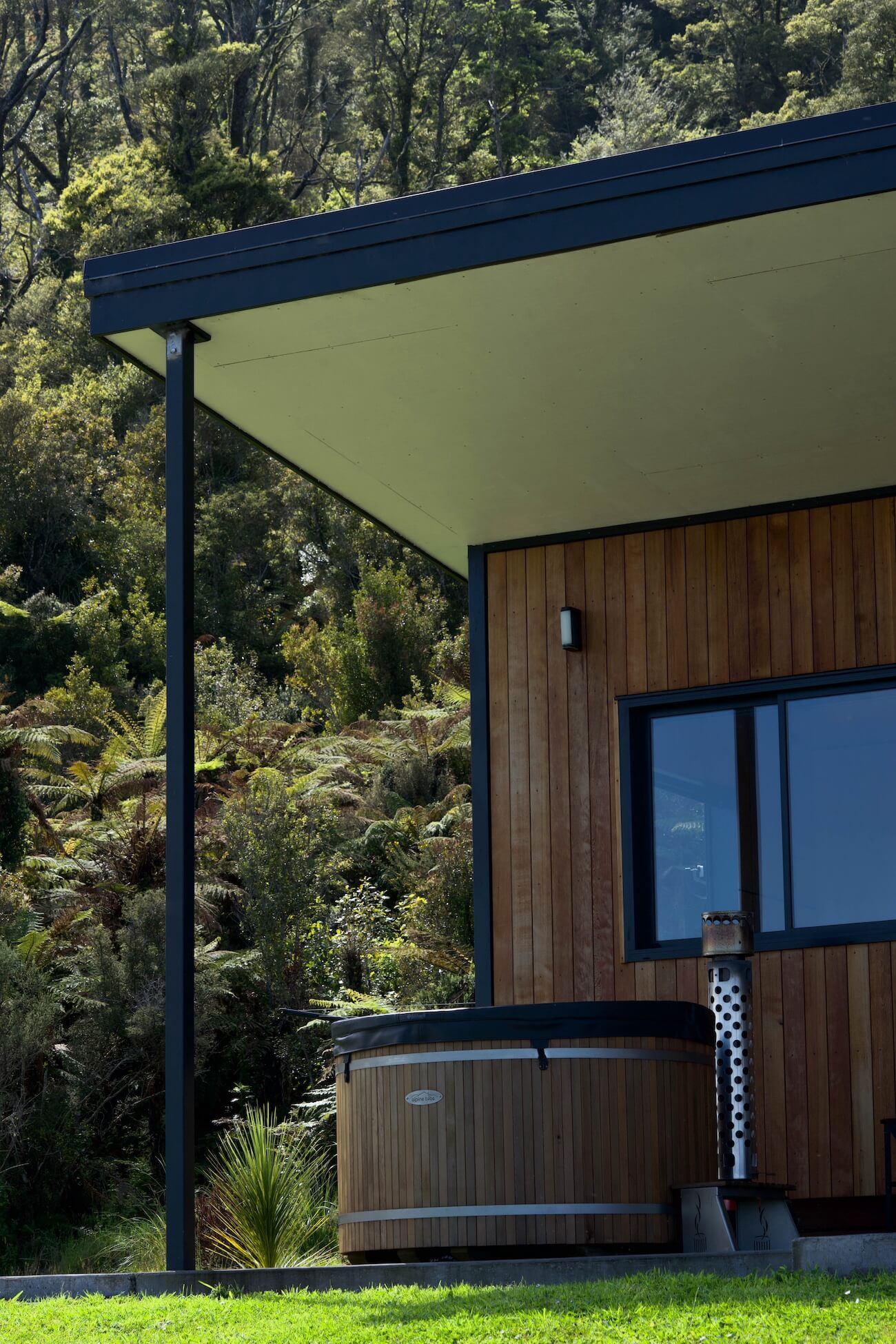 A modern wooden building in Karamea boasts large eaves, a window, and a hot tub on a lawn, all surrounded by dense greenery.