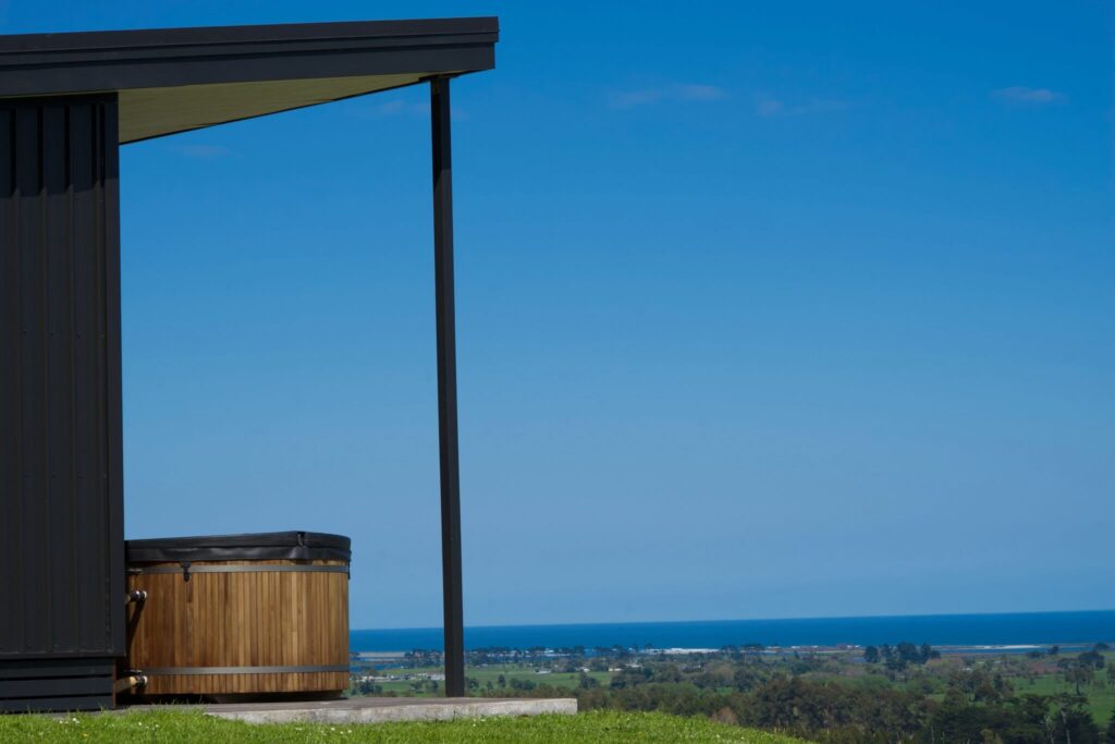 Modern house with a hot tub on a patio overlooking the lush green landscape and ocean near Karamea, all under a clear blue sky.