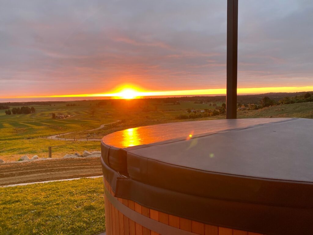 A panoramic view of a Karamea sunset over lush green fields, seen from the edge of a covered hot tub.