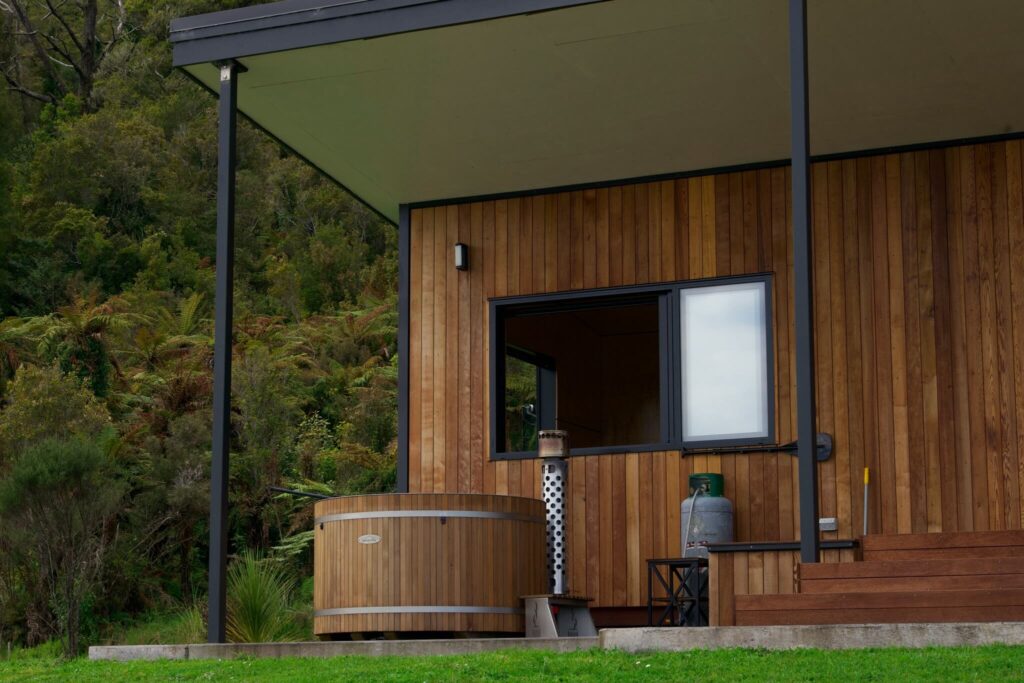 Nestled in Karamea, this wooden cabin boasts a small deck with a round hot tub, a metal heater, and a gas cylinder. Surrounded by lush greenery, it's the perfect retreat.