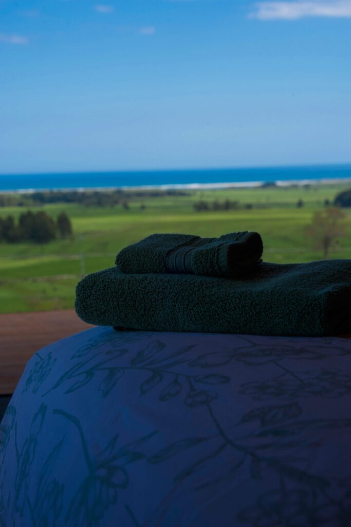 Folded green towels rest on a table, enhanced by the scenic beauty of Karamea’s grassy fields meeting the blue ocean in the background.