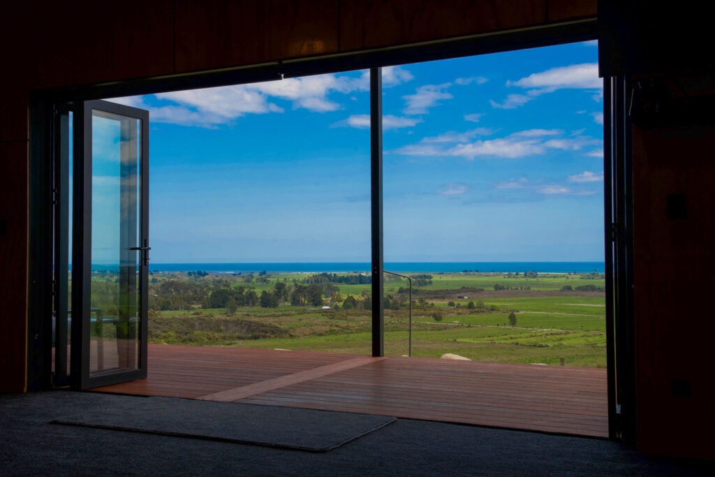 Open glass doors reveal a stunning view of Karamea's green fields and the ocean under a blue sky.