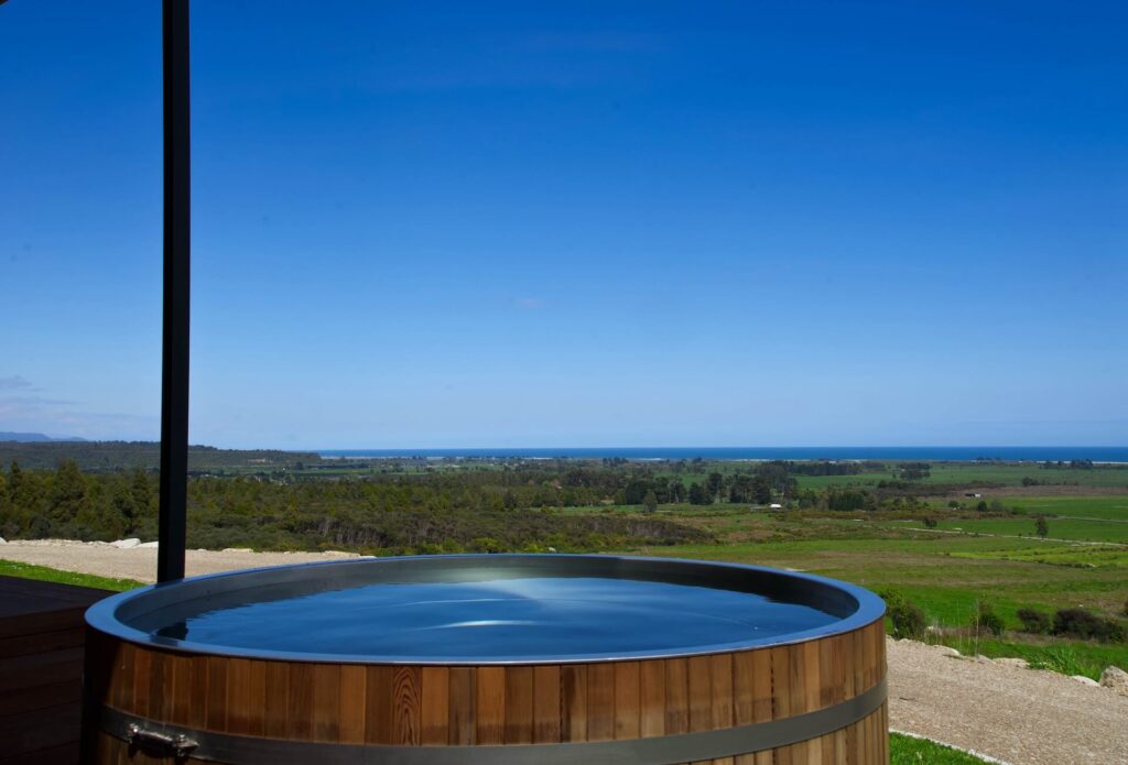 Nestled in the heart of Karamea, this wooden hot tub offers pristine clear water and overlooks a vast green landscape with the distant sea shimmering under a clear blue sky.